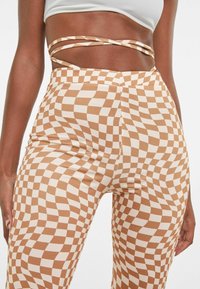 Legginsy w kratkę w ciepłym odcieniu brązu i kremu; dopasowane, o gładkiej fakturze i asymetrycznym detalu w pasie.