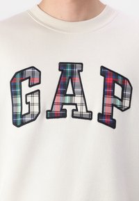 Kremna trenerka z velikimi izvezenimi črkami "GAP" v karirastem vzorcu, ki vključuje barve, kot so rdeča, zelena in Modra.