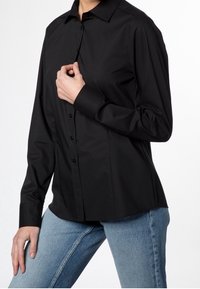 Personne portant une chemise noire cintrée à boutons avec des manches longues et un jean bleu, posant avec une main près du col.