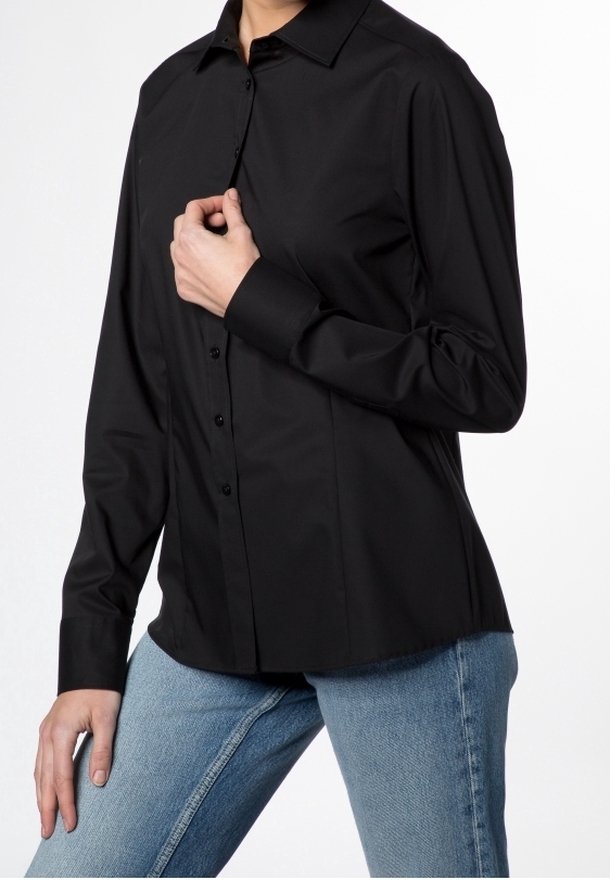 Personne portant une chemise noire cintrée à boutons avec des manches longues et un jean bleu, posant avec une main près du col.