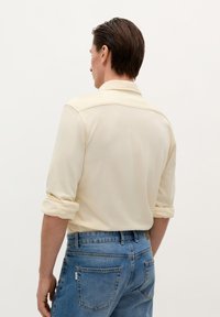 Light beige long-sleeve shirt with rolled-up sleeves, paired with mid-blue denim jeans. The shirt features a simple collar and soft texture.