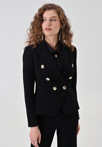 Blazer noir à double boutonnage en motif chevrons, avec des boutons dorés, des poches latérales, une coupe ajustée et des épaules structurées.