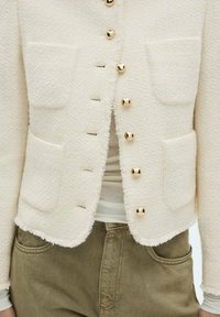Cream textured jacket featuring four front pockets, gold-tone buttons, and frayed edges, paired with a white top and olive-green pants.