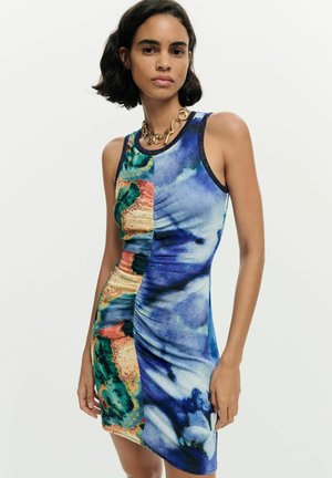 Žena v bezrukávových priliehavých šatách s abstraktnými viacfarebnými a modrými vzormi, stojaca na bielom pozadí.