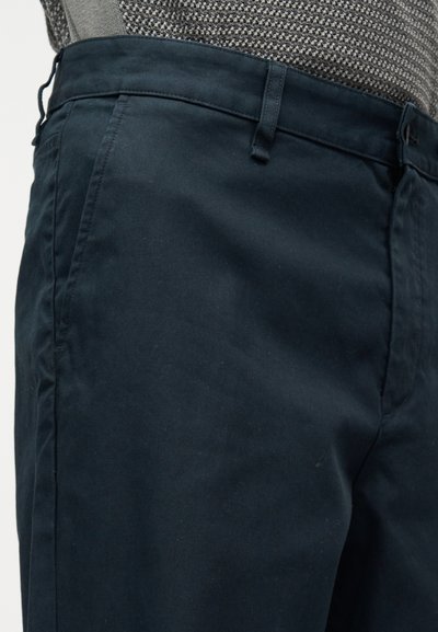 Άνδρας που φοράει σκούρο μπλε παντελόνι και γκρι ζακάρ πουλόβερ, πλάγια όψη με έμφαση στη μέση και την περιοχή του άνω μέρους του ποδιού.