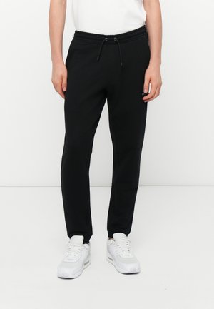 Sorte joggebukser med elastisk talje og snøre. Har to sidelommer og en tapered ben-design. Sammen med hvide sportssko.