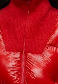Gros plan d'une veste rouge avec une partie supérieure en tricot et une partie inférieure en tissu matelassé brillant, avec une fermeture éclair centrale.