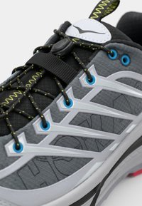 Chaussure de course avec un dessus gris, accents noirs et détails réfléchissants blancs. Présente un matériau texturé, des œillets bleus contrastants et des lacets jaunes.