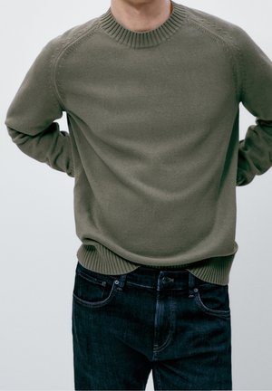 Mann mit hinter dem Rücken verschränkten Händen trägt einen olivgrünen Strickpullover und dunkelblaue Jeans vor neutralem Hintergrund.