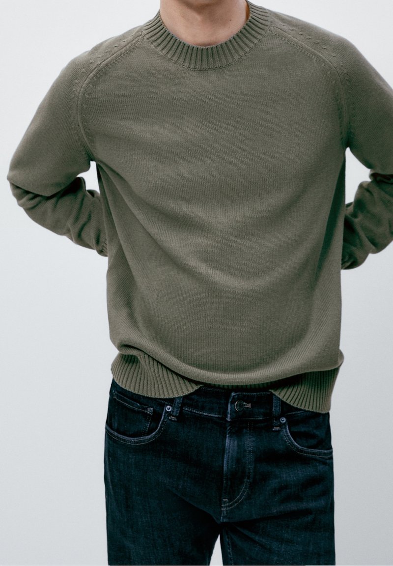 Homme les mains dans le dos, portant un pull en maille vert olive et un jean bleu foncé sur un fond neutre.