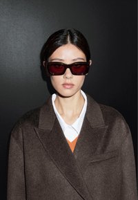 Brązowy wełniany płaszcz na białej koszuli z kołnierzykiem i pomarańczowym akcentem, w połączeniu z dużymi, ciemnoczerwonymi prostokątnymi okularami przeciwsłonecznymi.