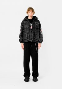 Chaqueta de plumas negra con múltiples bolsillos frontales, con cierre de cremallera y bordado floral. Combinada con pantalones negros de corte holgado y zapatos texturizados.