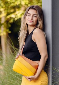 Žlutá a oranžová kožená kabelka přes tělo s diagonálním blokováním barev, hladkým povrchem a stříbrným zipem. Minimalistický design s detailem loga.