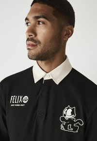 Schwarzes Polo-Shirt mit weißem Kragen. Auf der Vorderseite ist ein grafisches Design mit einer Cartoon-Katze und Text zu sehen. Aus Baumwolle, mit gerippter Textur.
