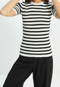 Schwarz-weiß gestreiftes Strickoberteil mit gerippter Struktur, rundem Ausschnitt und kurzen Ärmeln, kombiniert mit schwarzer Hose.