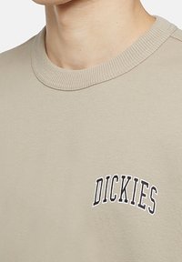 Felpa beige chiaro con collo rotondo a coste e logo ricamato "DICKIES" in nero. Tessuto morbido con vestibilità comoda.