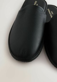 Black leather slippers with a smooth texture, rounded shape, and soft upper. Features a cushioned footbed and rubber outsole for grip.
