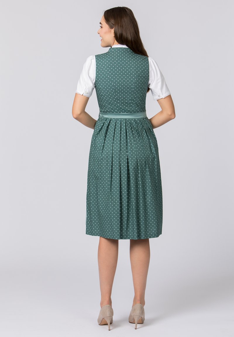 Green polka dot dress with a fitted bodice and pleats at the skirt; paired with a white blouse. Features a satin waistband and neutral shoes.