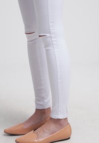 Białe podarte wąskie dżinsy noszone z beżowymi balerinami w szpic, ukazujące dolne części nóg i stopy na jednolitym tle.