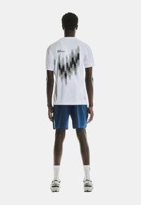 T-shirt blanc à manches courtes présentant un design graphique noir au dos, associé à un short bleu et des chaussettes blanches, porté avec des baskets.