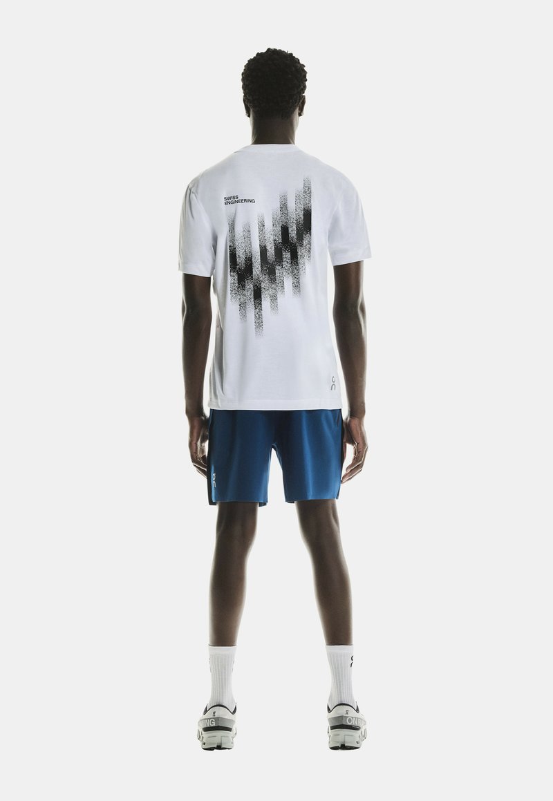 T-shirt blanc à manches courtes présentant un design graphique noir au dos, associé à un short bleu et des chaussettes blanches, porté avec des baskets.
