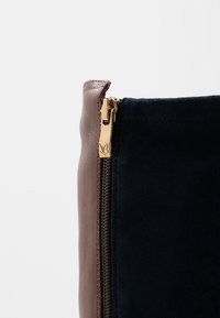 Bolso bandolera de cuero marrón y ante azul marino con cremallera dorada y detalles en oro; presenta una textura suave y materiales contrastantes.
