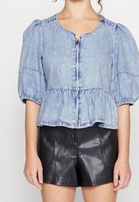 Ljust blå puffärmad denimblus med framzipper, tillsammans med högmidjade svarta lädershorts med knappar och fickor.