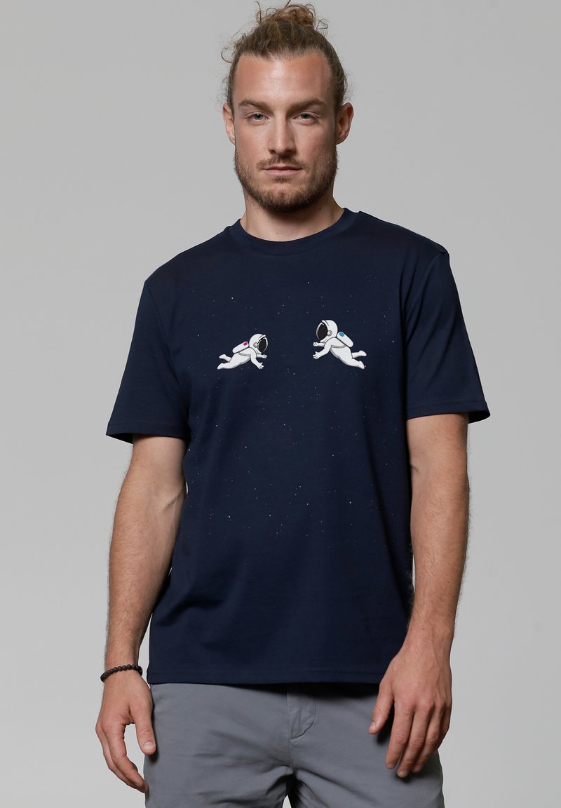 Navyblaue Baumwoll-T-Shirt mit zwei weißen Astronauten-Grafiken, einem sternenübersäten Hintergrund und subtiler Textur im Stoff. Kurze Ärmel, Rundhals.