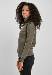 Giovane donna con capelli ricci che indossa una felpa zip-up verde oliva e jeans neri, in piedi di profilo su uno sfondo grigio uniforme.