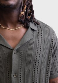 Olivgrünes, gestricktes Hemd mit Knopfleiste, kurzen Ärmeln und vertikalem Streifenmuster mit kleinen Aussparungen. Sichtbare goldene Kettenhalskette.