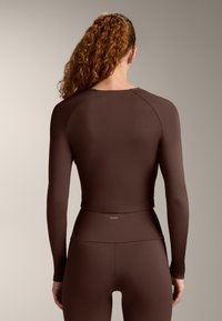 Vrouw met krullend rood haar in een hoge paardenstaart, gekleed in een nauwsluitend, bruin sportief shirt met lange mouwen en bijpassende hoog getailleerde legging, met de rug naar de camera toegekeerd.