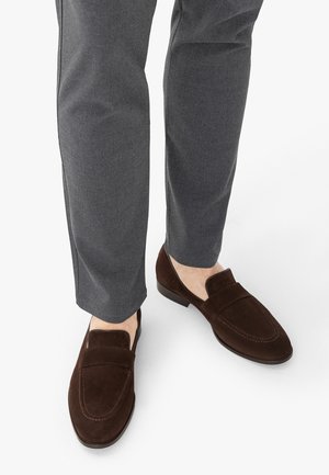 Mann trägt graue Hose und dunkelbraune Wildleder-Loafer vor weißem Hintergrund, nur Füße und Unterschenkel sichtbar.