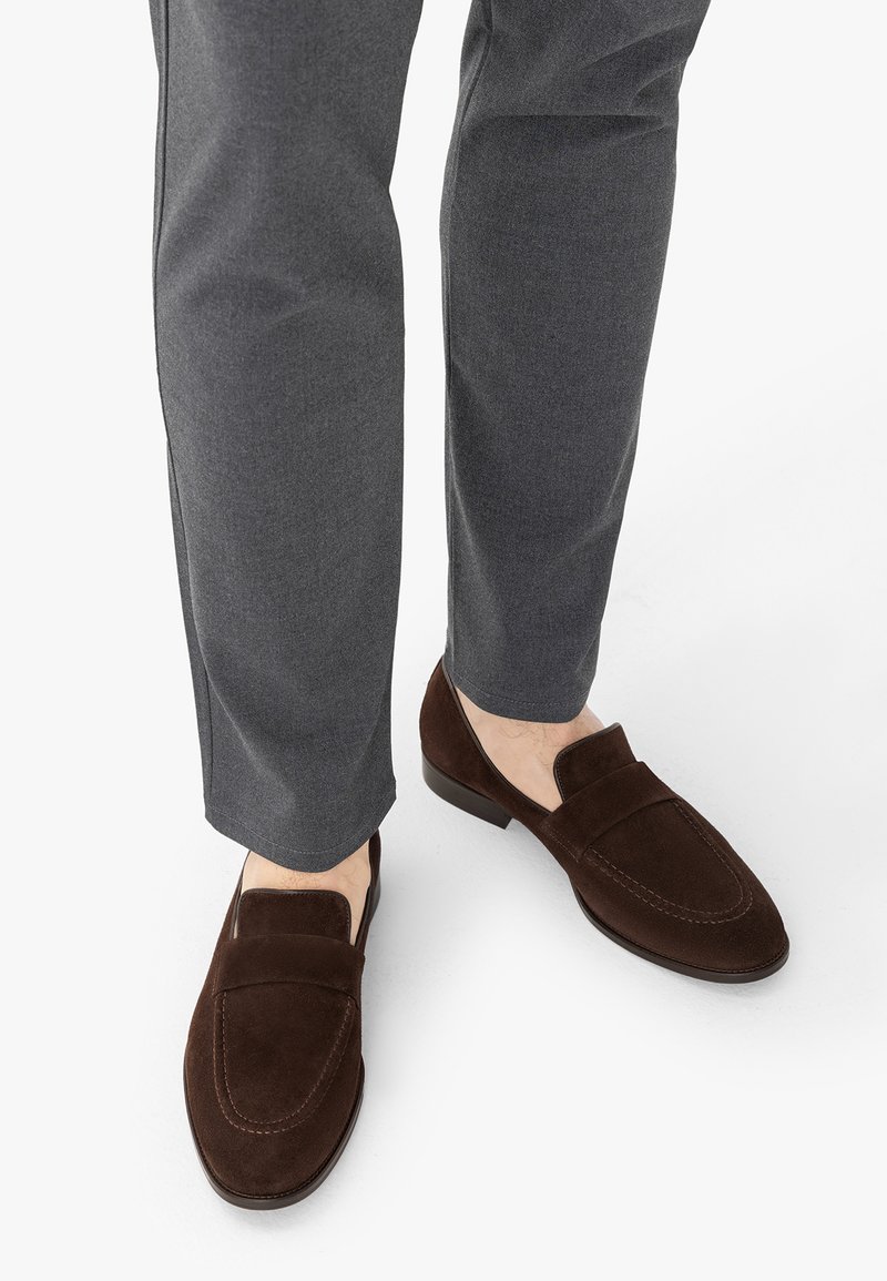 Homme portant un pantalon gris et des mocassins en daim marron foncé sur un fond blanc, ne montrant que les pieds et le bas des jambes.