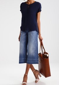 Blusa blu navy con dettaglio a keyhole, jeans blu a gamba larga con orlo sfilacciato, décolleté beige con cinturino alla caviglia e borsa a tracolla in pelle marrone.