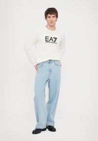 Hvit langermet skjorte med svart EA7-logo, kombinert med lyseblå vide jeans og svarte sko. Modellen står med hendene på hoftene.