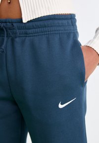 Tummansiniset collegehousut, jotka on valmistettu pehmeästä kankaasta. Niissä on joustava vyötärö ja sivutaskut; vasemmassa lahkeessa on valkoinen Nike-logo.