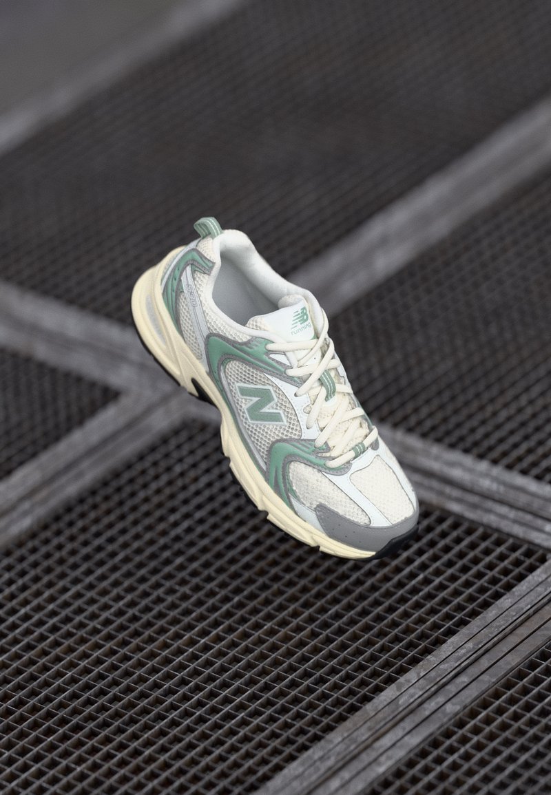 Fehér és mentazöld New Balance futócipő egy fekete fémrács felület fölött lebeg.