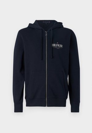 Marineblauwe zip-up hoodie van zacht materiaal. Beschikt over een kangoeroe pocket en logodetails op de linkerborst. Inclusief trekkoord capuchon.