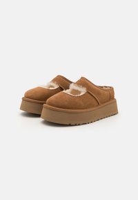 Brown suede clogs with a thick, textured sole and inner lining of soft white fur. Design features an open back and minimalist stitching.