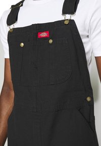 Bretelle in denim nero con hardware dorato, spalline regolabili, tasca frontale e patch con logo sul petto. Presenta un classico fit rilassato.