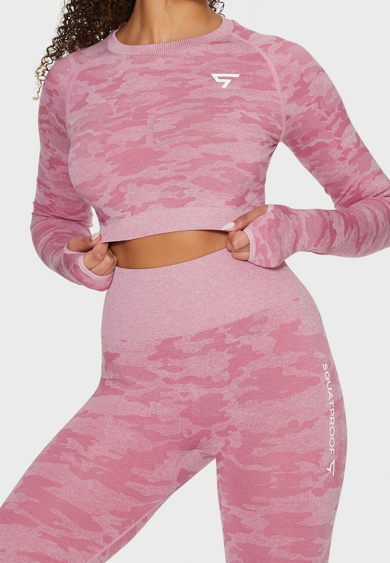 Femme portant un crop top manches longues camouflage rose et des leggings taille haute avec des trous pour les pouces et des logos Fitness sur la poitrine et la cuisse.