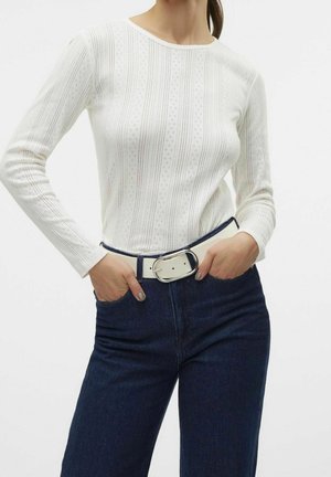 Femme portant un haut à manches longues blanc à motifs, jean bleu foncé, ceinture blanche avec une grande boucle argentée, mains dans les poches.