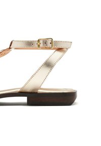 Sandalia plana con tiras metálicas en dorado rosa, con un detalle de hebilla. Parte superior de cuero texturizado con una suela de goma marrón y lisa.