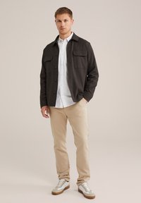 Tumši pelēks pogājams žakets ar divām krūšu kabatām, valkāts virs baltas krekla ar apkakli, saskaņots ar bežām biksēm un baltām tenisēm.