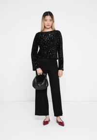 Top lung cu maneci lungi din sequinuri negre, cu decolteu în formă de barcă, asortat cu pantaloni largi negri. Modelul ține o geantă neagră și poartă pantofi roșii cu vârf ascuțit.