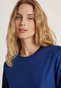 Blue cotton t-shirt with a round neckline and long sleeves, featuring a smooth texture. The model has wavy hair and a neutral expression.