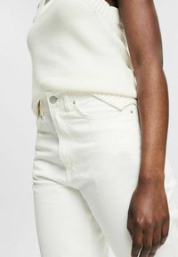 Top lavorato a maglia bianco con scollatura a girocollo abbinato a jeans a vita alta di colore bianco sporco, dotati di tasche laterali e dettagli con bottoni metallici.