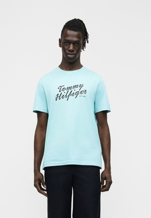 Giovane uomo con dreadlocks che indossa una maglietta azzurro chiaro Tommy Hilfiger e pantaloni scuri, in piedi davanti a uno sfondo bianco uniforme.