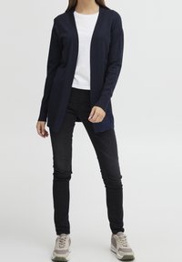 Marinblå stickad cardigan med öppen front, långa ärmar och ribbat detaljer. Kombineras med en vit topp och svarta jeans. Sneakers med en beige/grå färgkombination.