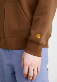 Sudadera de algodón marrón con puños acanalados y un logotipo bordado en dorado, diseñada con un corte relajado y un bolsillo tipo canguro.